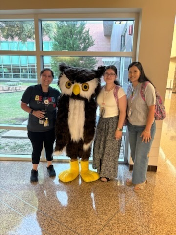 Four people stand in a hallway; one wears an owl mascot costume. They are near a window with a grassy view.