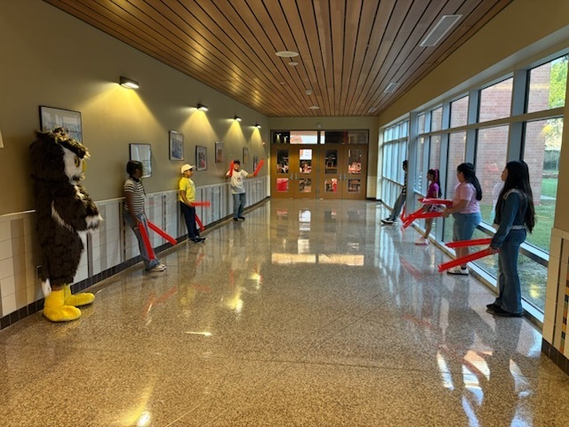 Children hold red sticks and stand in a hallway with a mascot and large windows.
