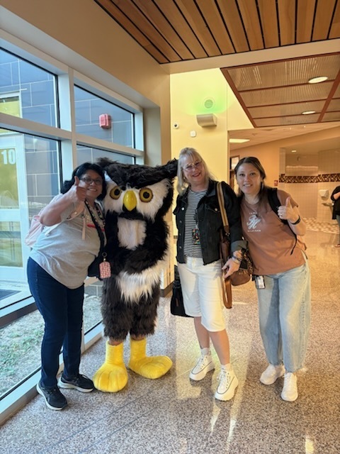 Four individuals stand in a hallway; two women pose with an owl mascot and a large building window behind them.