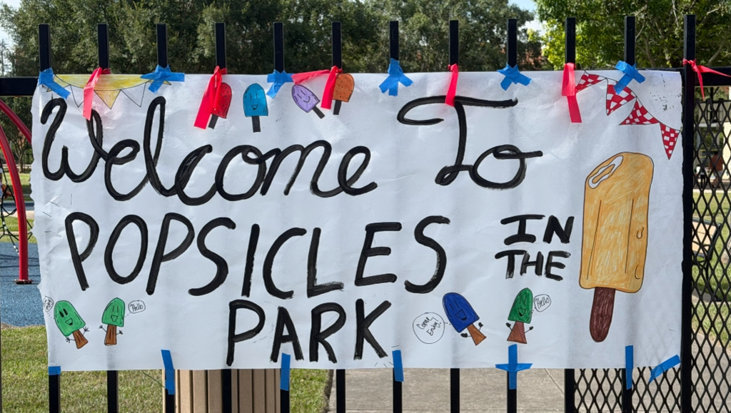 A banner with colorful popsicle drawings reads "Welcome To Popsicles In The Park." It hangs on a black fence.