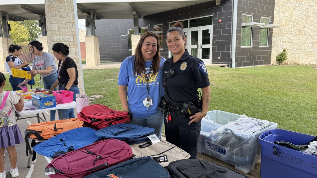 Two women stand by a table with backpacks and bags in front of a building with large windows.
