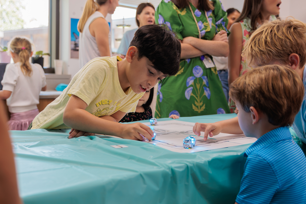 Kids interact with small, colorful objects on a table covered with a teal cloth, in a room with several adults watching.