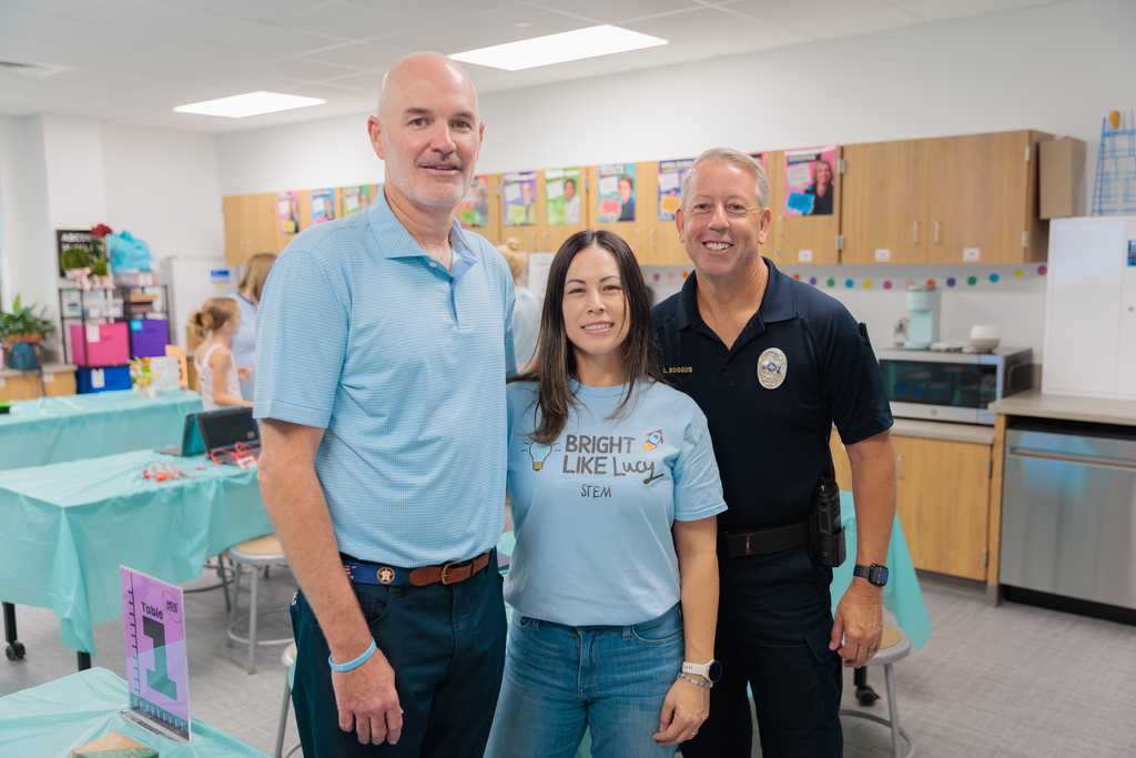 Three people in a room with tables, cabinets, and a microwave. The two men wear blue shirts, and the woman wears a light blue shirt.