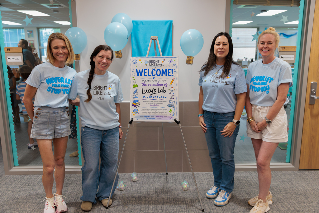 Four women stand together in a room with a welcome poster, wearing blue shirts. Balloons and a door are behind them.