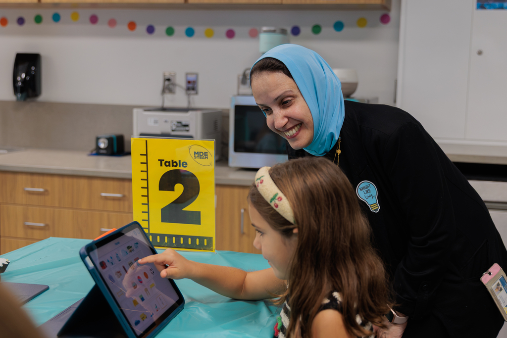A smiling woman and a child using an iPad. The table has a blue cover. Behind them, a microwave and coffee maker.