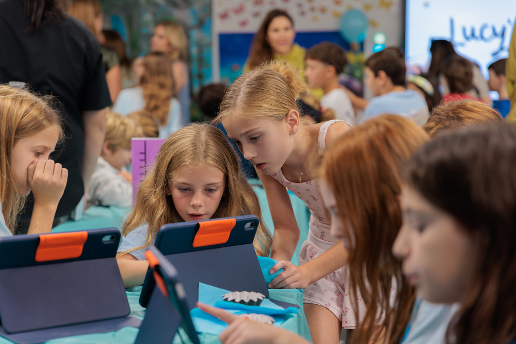 Children in a classroom setting are using laptops and tablets; they are seated in a semi-circle.