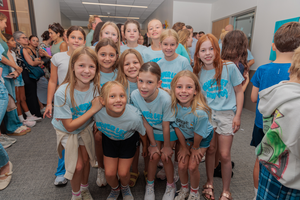 Group of young girls in matching blue shirts posing for a photo with others in a hallway.