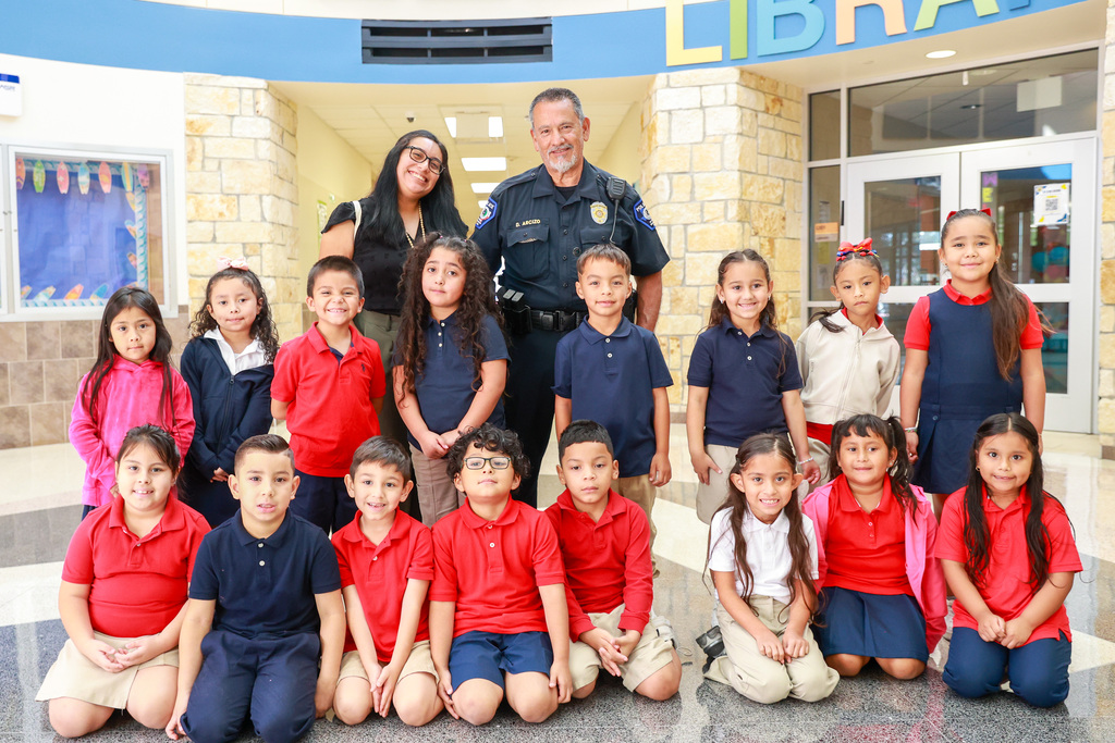 School children in matching shirts, some red, some blue, posing with a uniformed police officer inside a building.