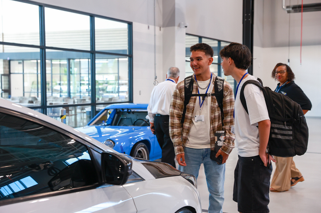 Two young men in a car showroom, one wearing a backpack. A blue car is parked behind them.