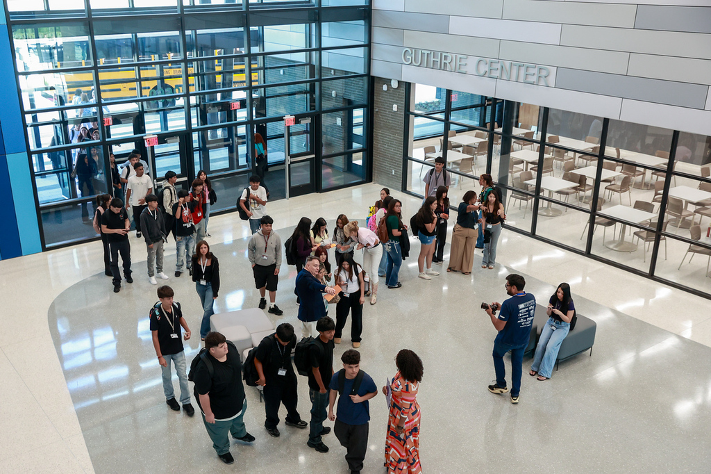 A room with glass walls, large tables, and a bus visible through the windows. People are gathered, some in conversation.