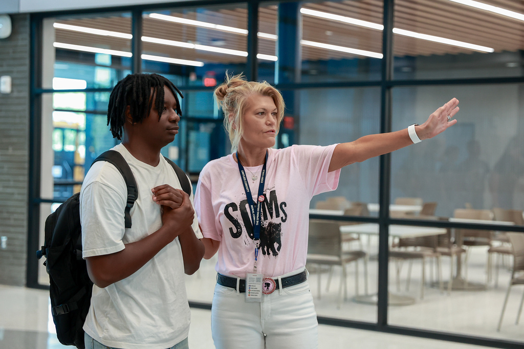 A man and a woman are standing and talking in a cafeteria with tables, chairs, and glass walls.