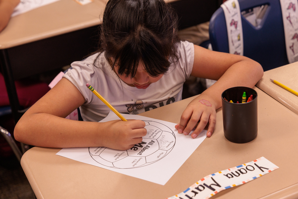 Child seated at a desk in a classroom, coloring a large circle with a pencil. Nearby is a cup with colored pencils.