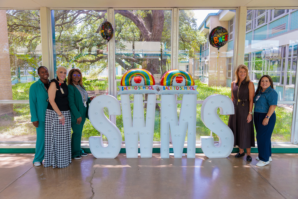 Five women stand behind a white sign reading SWMS, with a tree, balloons, and a building in the background.