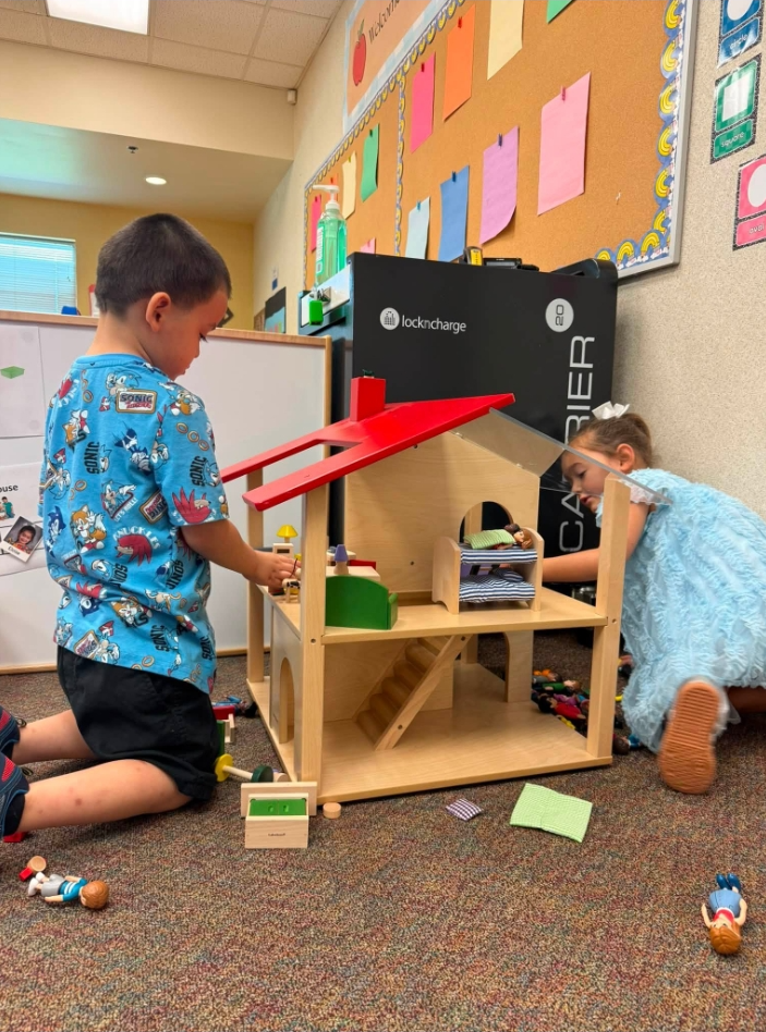 Two children play with a toy house on a carpeted floor. A child in a blue shirt and black shorts.