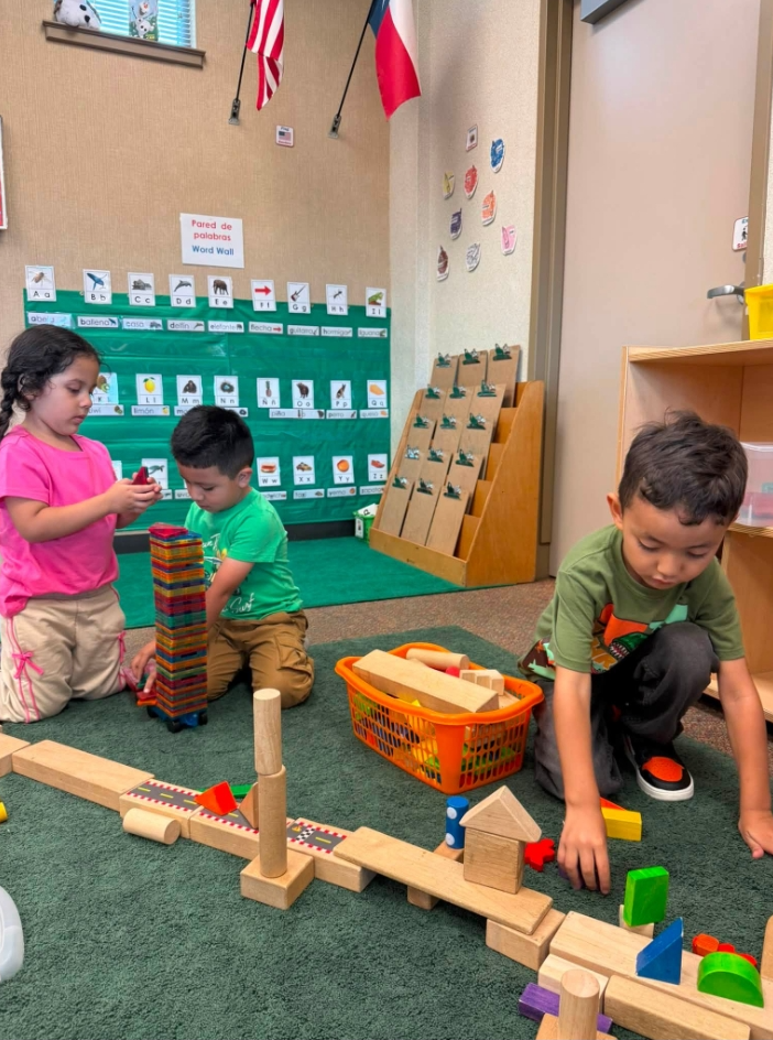 Children playing with building blocks on a green rug in a classroom with flags.