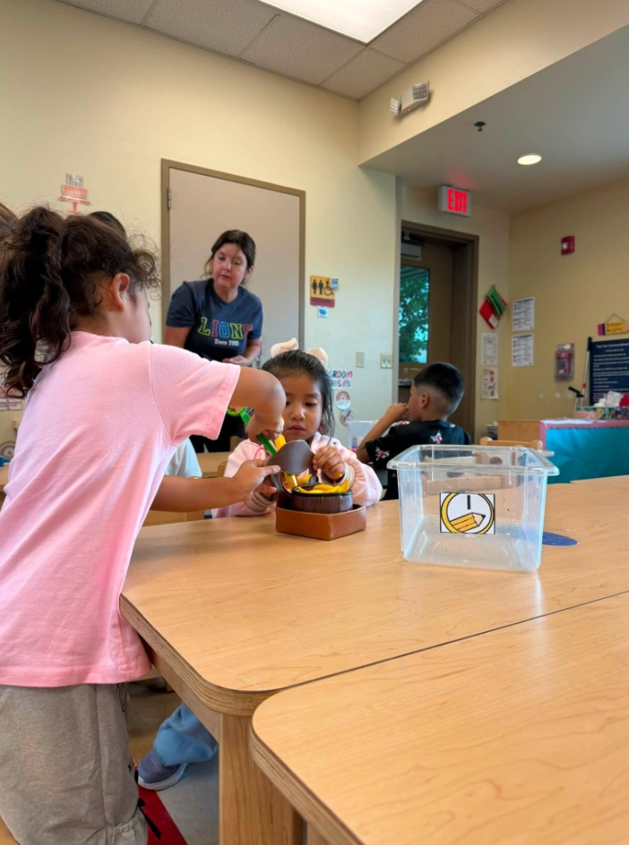Kids in a classroom work on a cake together at a table. A woman stands behind them.
