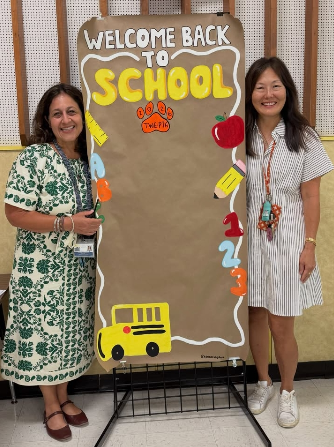 Two women stand next to a school sign. It says "Welcome Back to School" with school objects. The background is a wall.