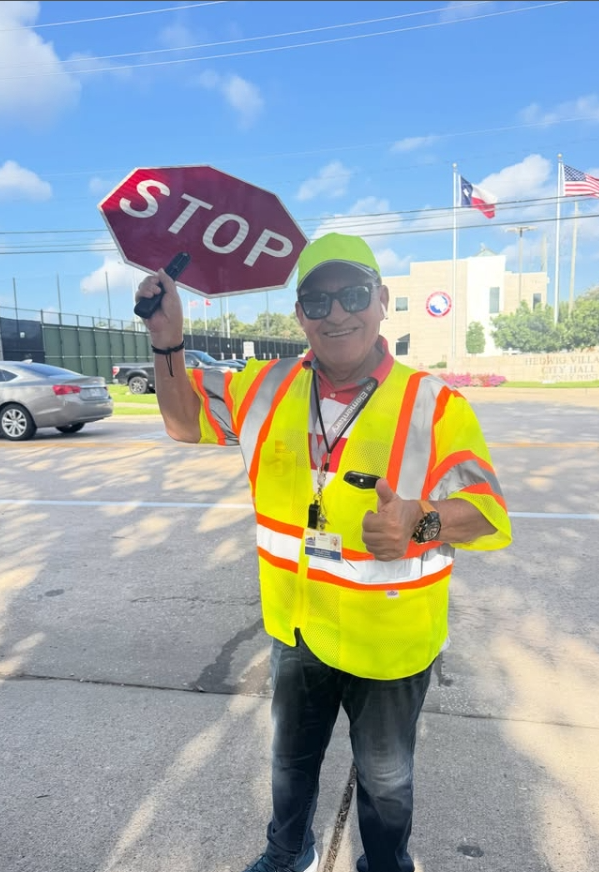 A man in a yellow vest holds a stop sign and gives a thumbs-up gesture. Behind him, there are cars, a building, and flags.
