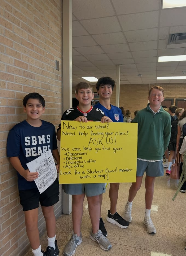 Four boys stand in a hallway, one holding a yellow sign with words "ASK US!" and another holding a white sign.