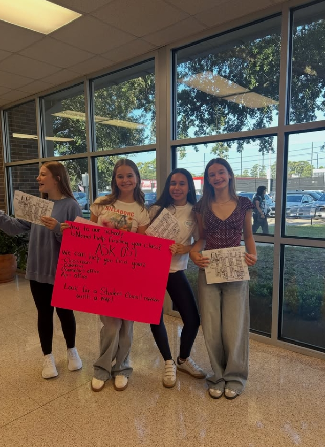 Four young women holding signs in a school hallway. Two of them hold pink signs. The glass windows behind them reveal a parking lot with cars and trees.
