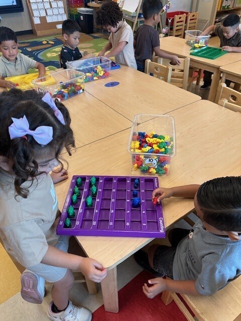 Several young children seated around tables in a classroom. A girl and boy are playing a board game with colored cubes.