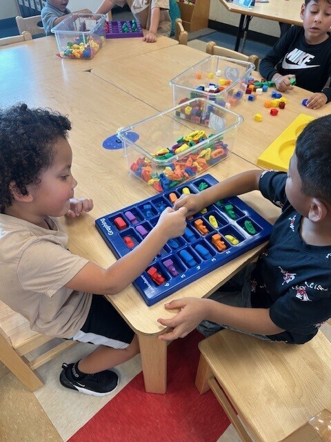 Two children play with a blue tray of plastic shapes at a wooden table. Other children work at tables behind them.