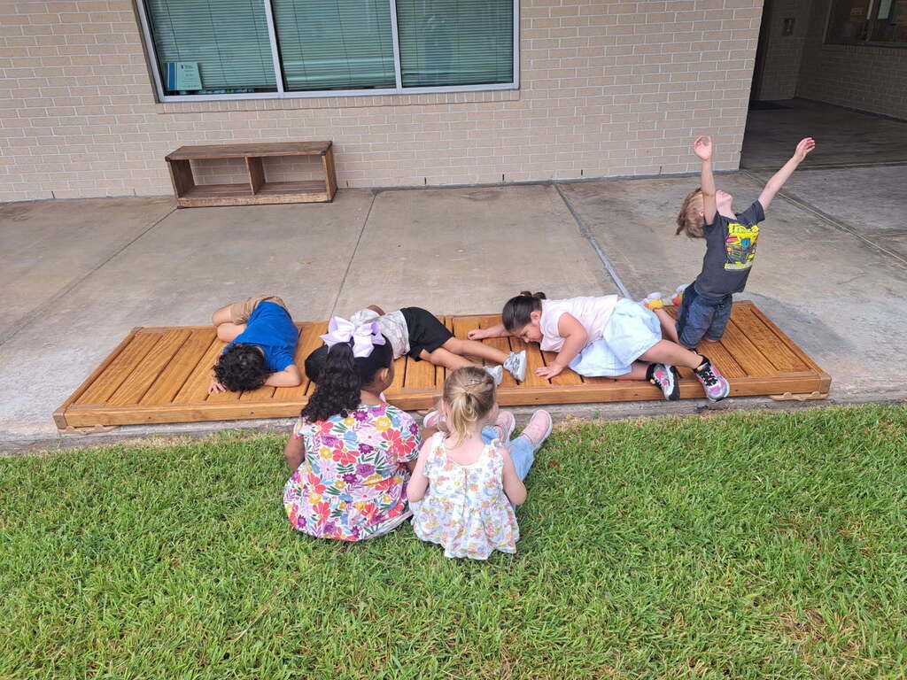 Children seated and standing on a wooden ramp outside a house, one with arms raised.