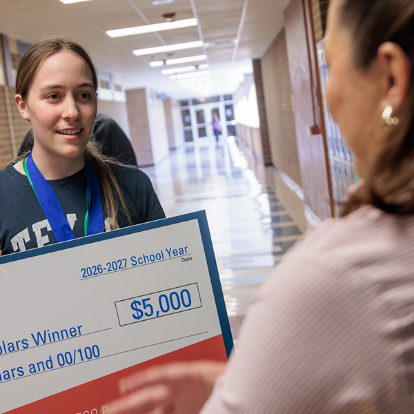A girl holding a check for $5,000. She stands in a hallway, smiling and facing a woman.