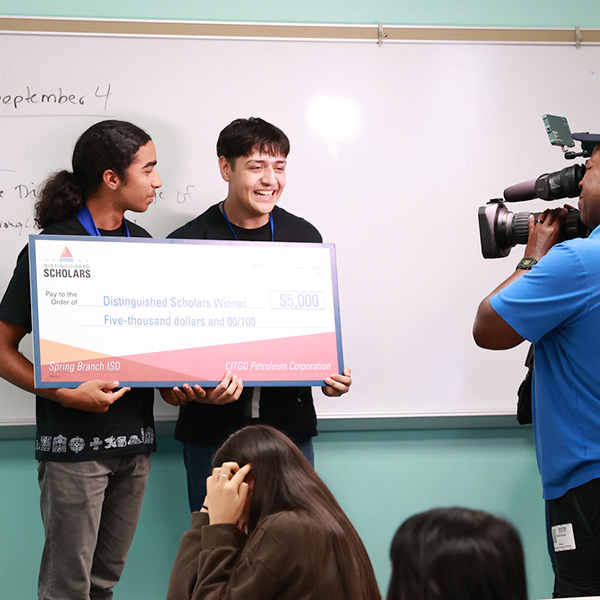 Two men hold a large check, smiling. A cameraman films them in front of a whiteboard.