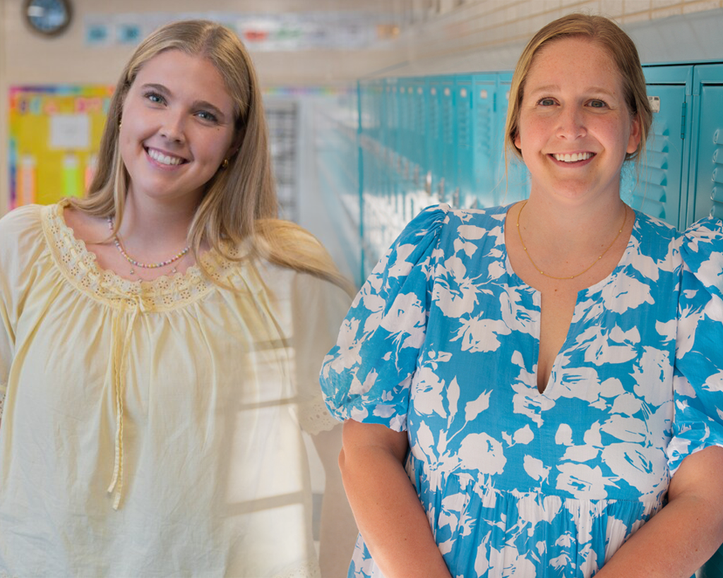Two women, one in a yellow dress and the other in a blue and white floral dress, smiling and standing in a school hallway.