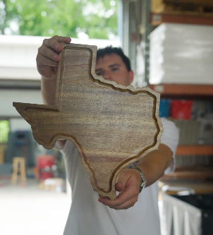 A man in a white shirt holds a Texas-shaped wooden cutting board in a storage area.