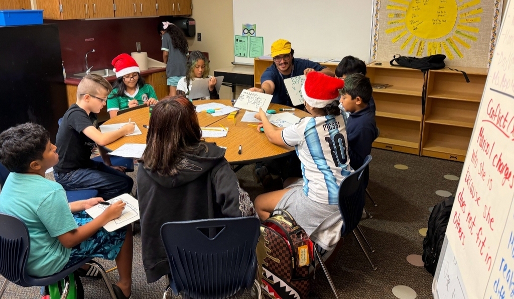 A group of students wearing hats are sitting around a table and discussing in a classroom.