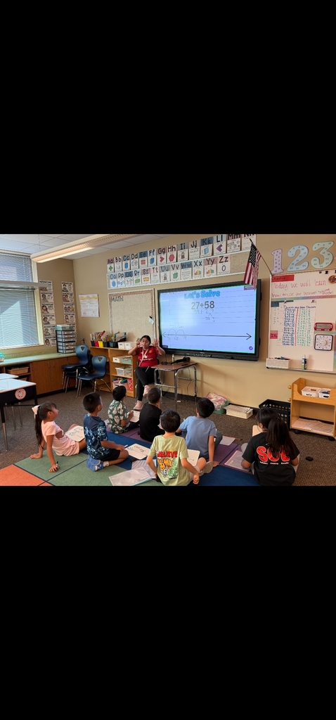 Classroom with students sitting on the floor. A teacher stands before a screen. Bulletin boards and a flag are visible in the background.