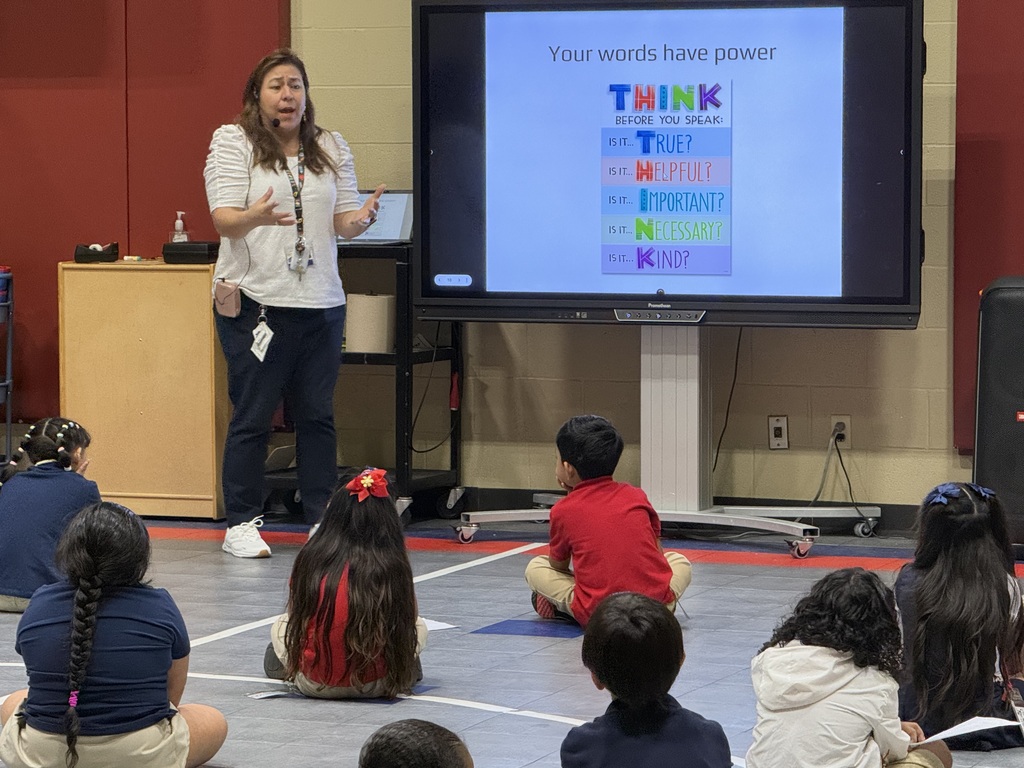 A teacher presents a lesson in a classroom. The screen displays "Your words have power." Students are seated on the floor.