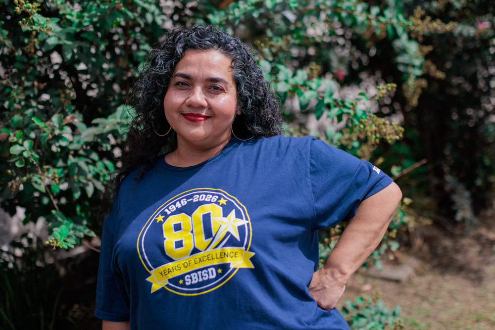 A woman smiles and poses outside near greenery, wearing a navy blue shirt with "80" and a star logo.