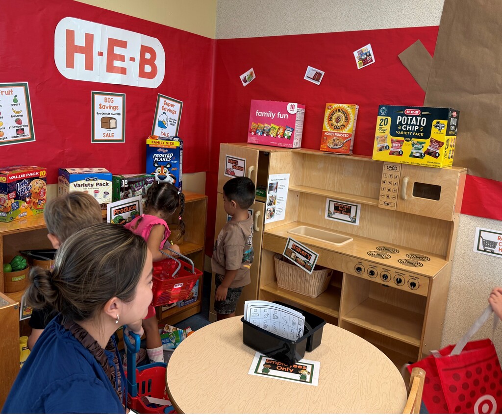 A room with red walls and a sign reading H-E-B. Children explore shelves with toys.