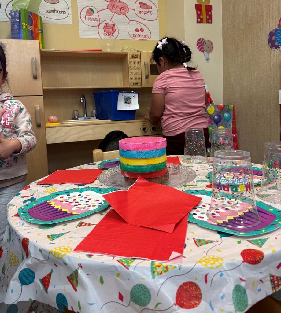 Two children in a classroom setting. One child is arranging a colorful cake and napkins on a table.