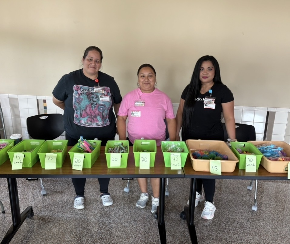 Three women in casual clothes stand behind a table with green bins. One wears a pink shirt, while the others wear black.