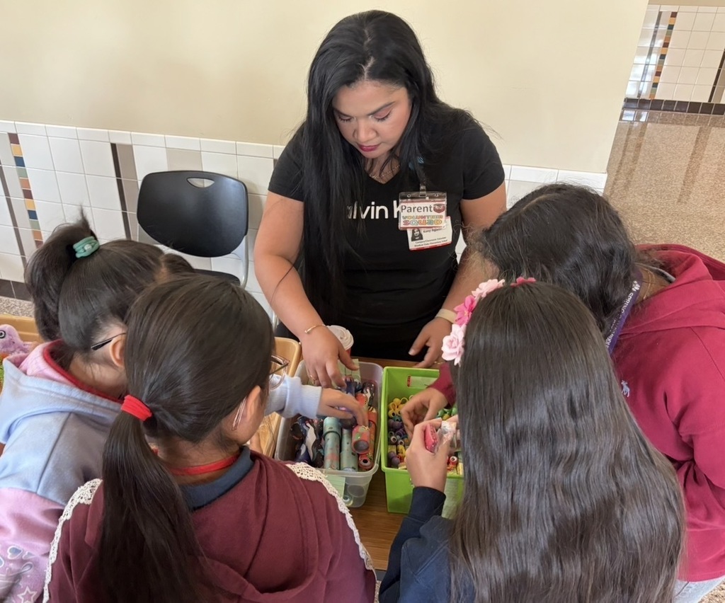 Woman leading young students in a classroom, seated around a table with small items, tiled wall in background.
