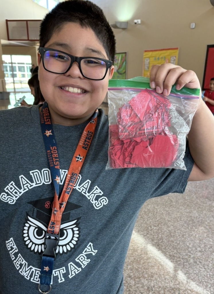 A child holds a plastic bag containing heart-shaped objects. The child wears a gray T-shirt and glasses. The background shows a hallway.