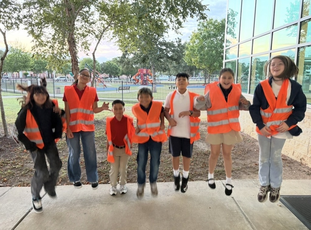 A group of children in orange safety vests pose on a sidewalk. Trees and a building are in the background.