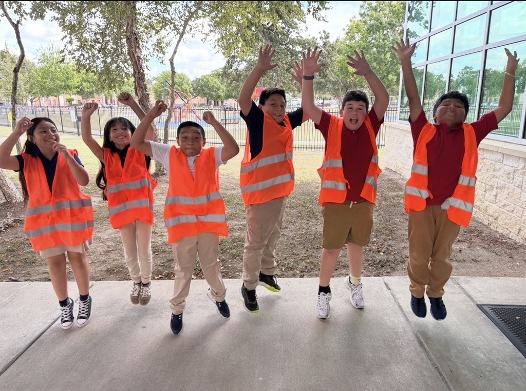 Kids wearing orange vests and sneakers pose in front of a building with trees. They jump with hands raised.
