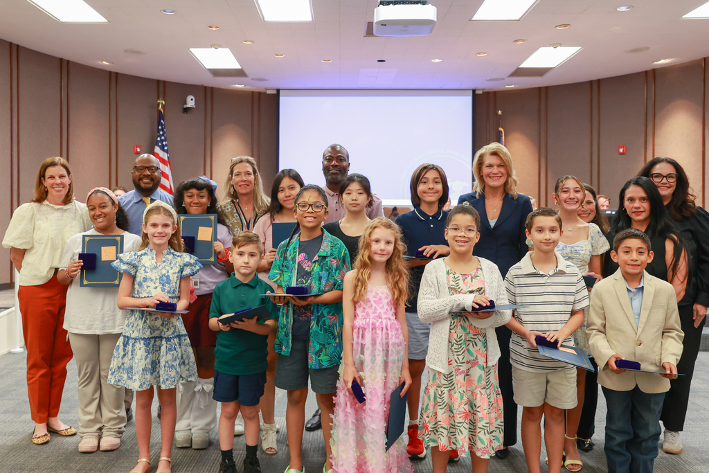 A group of adults and children holding awards stand in a circle. A woman stands in the middle.