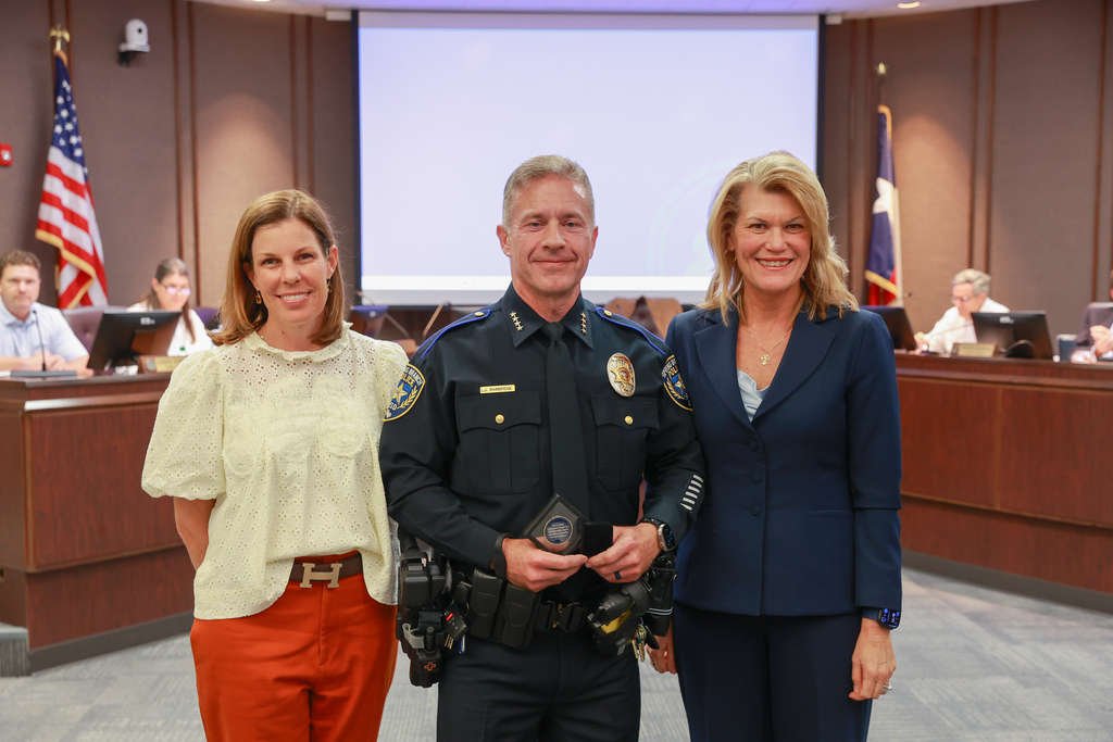 Three people stand in a room; a man in police uniform holds an award, flanked by two women. A flag is visible in the background.