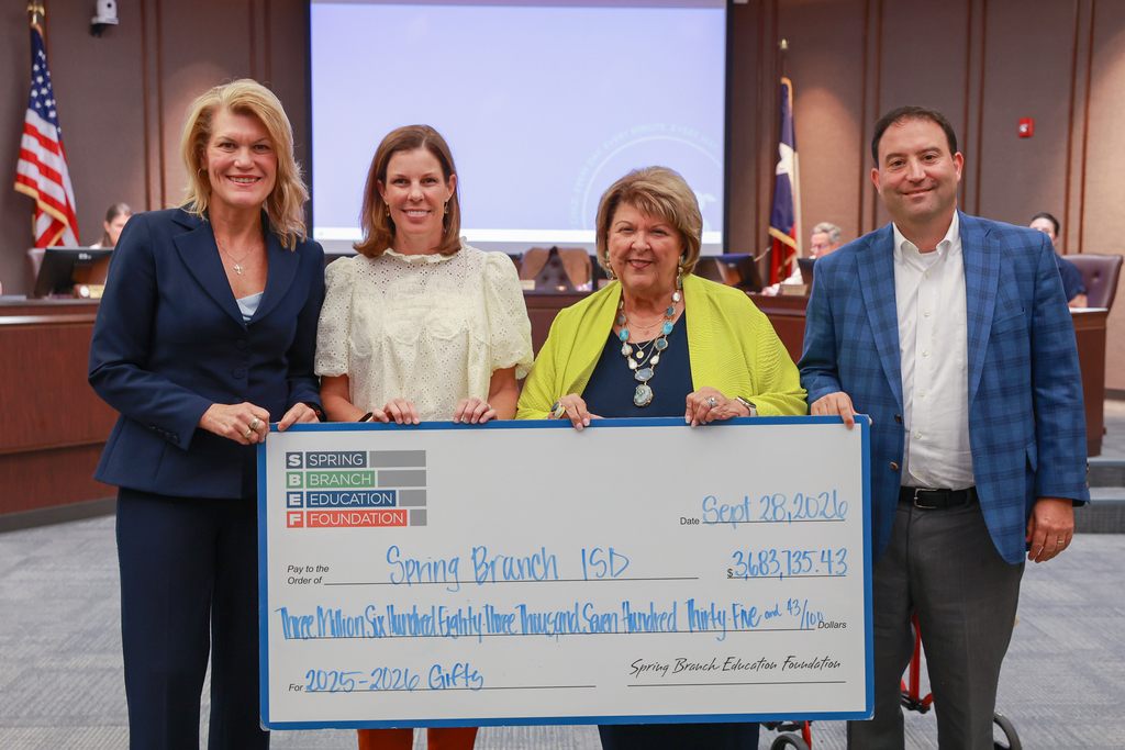 Four people hold a large check. Two women on the ends wear suits; one wears a dress. A flag and chairs in the background.
