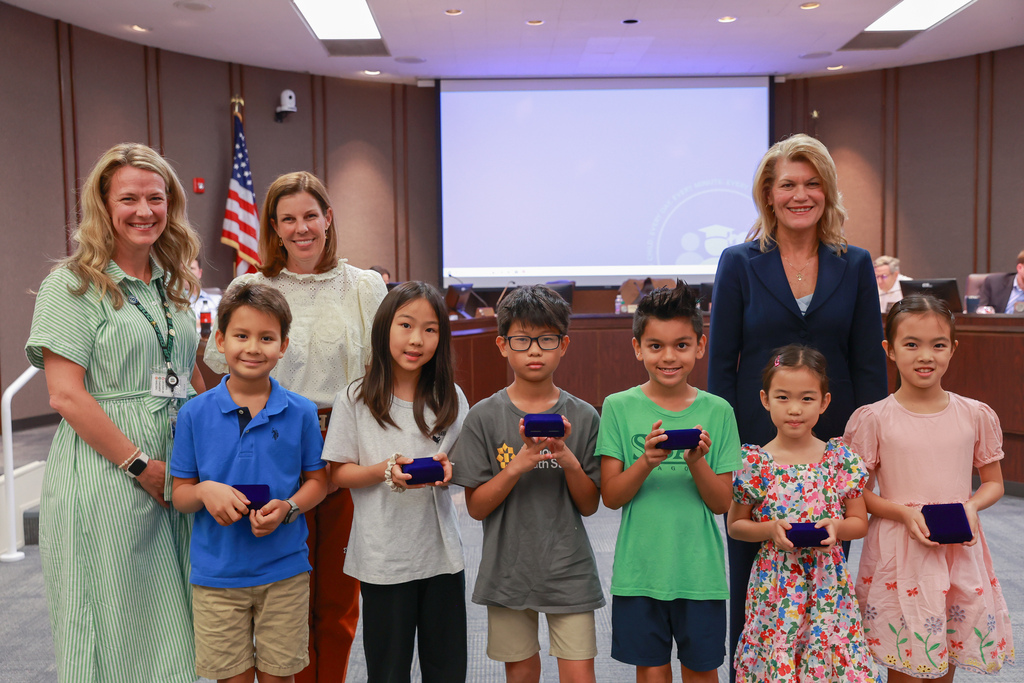 Several adults and children holding awards stand together in a conference room. Two women stand at the sides.