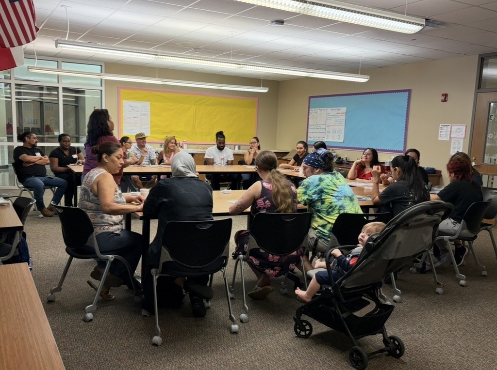 A group of people seated around tables in a classroom, with a baby in a stroller and an American flag visible.
