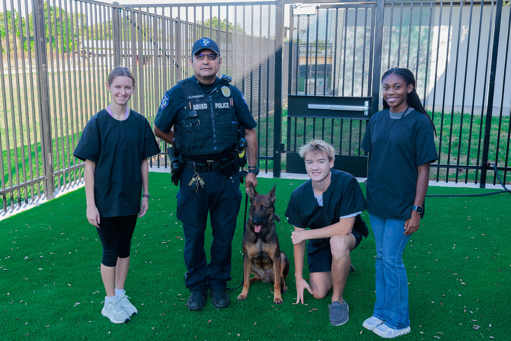 A group of people stand on artificial turf. A police officer with a dog, two teens, and a young woman are present.