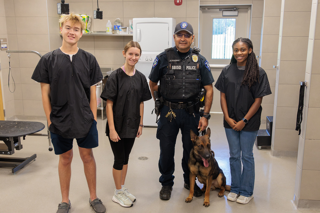 Five people stand in a room, including a police officer with a German Shepherd, a boy, and three girls.