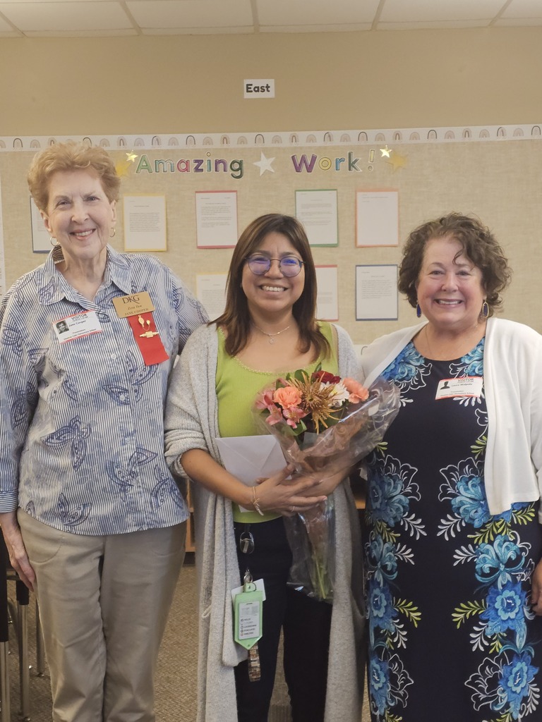 Three women stand together, smiling, in a room. The center woman holds a bouquet. A bulletin board with text is behind them.